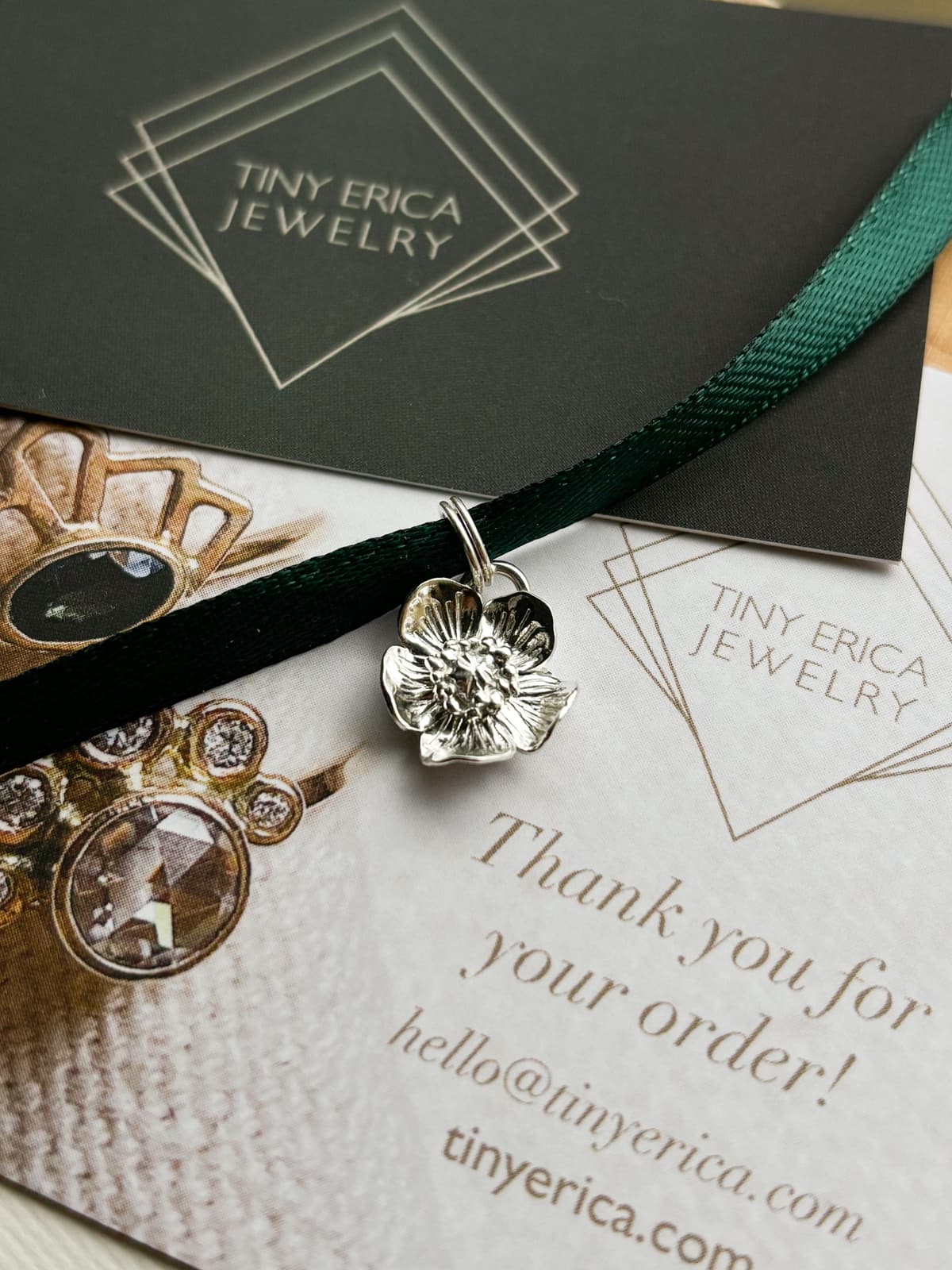 a sterling silver flower pendant on a loop wrapped around emerald-colored ribbon lying atop two Tiny Erica Jewelry business cards